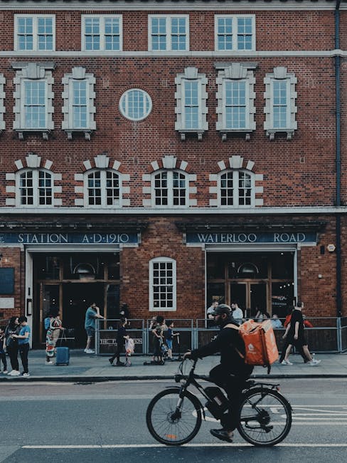 The image shows a brick building with large windows and a sign indicating Waterloo Station, with a crowd of people standing and walking along the sidewalk in front. In the foreground, a person dressed