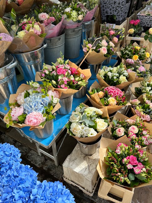 A display of freshly arranged flower bouquets featuring soft pink, white, and pastel blue blooms, including roses, hydrangeas, and other mixed floral varieties, wrapped in light brown craft paper and 